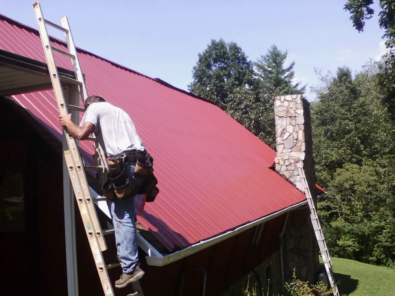 Roofer on a ladder beside a red metal roof and stone chimney — Soffit & Fascia Repair by Roofing Brothers in Stephens City